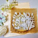 A floral wooden cutting or craft panel with layered white wood cutouts of flowers and leaves, displayed on a woven mat over burlap, with decorative flowers and a gold binder clip nearby.