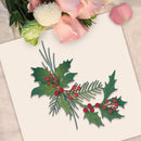 A decorative paper cutout of holly leaves and red berries arranged on a white card, with pink flowers placed around the edges as styling props.