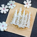 A white metal laser-cut die featuring four pine tree silhouettes, displayed on a gold wire stand over a brown kraft paper sheet, with decorative white flowers in the corners.