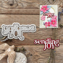 A flat-lay product presentation of a crafting die and a handmade greeting card on a rustic wooden background. The die is silver-colored metal with the words 'sending JOY' in an ornate script.