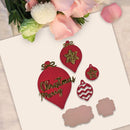 A flat-lay of red Christmas ornament-shaped craft pieces with gold glitter and gold lettering on a white card, styled with pink flowers at the top.