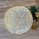 A set of silver metal cookie cutters with Christmas-themed designs, including ornaments, stars, and holiday phrases, arranged on a round woven straw mat over a wooden surface.