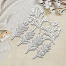A set of silver metal die-cut embellishments shaped like pine branches and leaves, arranged on a textured cream fabric with cotton stems as decorative props.
