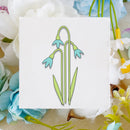 A square white card with a minimalist line drawing of snowdrops in light green and light blue, styled on a beige fabric background with white, blue, and yellow flowers.