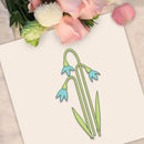 A decorative flat-lay showing a white card with a simple line drawing of three snowflake-like flowers in light green and light blue, surrounded by pink and white flowers on a light fabric surface.
