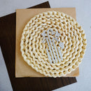 A round woven straw placemat in a light cream color, displayed on a square tan paper background with a silver metal monogram template placed in the center.