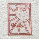 A handmade greeting card with a white background, coral-red geometric border, a layered heart design with small floral print, and the word 'Merci' in coral-red script, displayed on a white lace fabric background.