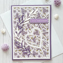 A handmade layered greeting card with a purple and white floral pattern, featuring a heart-shaped cutout, purple leafy embellishments, and a small banner reading 'JOYEUX ANNIVERSAIRE'. The card is displayed on a white wooden surface with a white envelope and decorative paper flowers.
