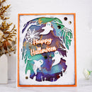 A square layered Halloween greeting card with an orange border, featuring white ghost cutouts over a purple and teal background with spiderwebs, bare trees, small crosses, and the words 'Happy Halloween' in orange script. The card is photographed on a white surface with decorative dried flowers at the side.