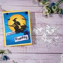 A Halloween-themed handmade card with a yellow border, blue striped background, a large orange moon, and a black witch silhouette on the front, shown beside a matching metal die-cut cutting mat on a light pink wood surface with small flowers around it.