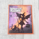 A square Halloween-themed card featuring a black silhouette of a witch with a broom, a cat, a spider, and a small black jack-o'-lantern, set against a purple and orange sunset background with a torn-paper 'HALLOWEEN' banner. The card has an orange border and is photographed on a light wood surface.