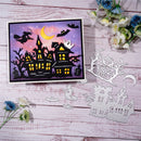 A Halloween-themed layered card or craft sheet displayed on a whitewashed wood surface, alongside matching silver die-cut paper cutouts of a haunted house, witch on a broomstick, ghost, moon, and tree. The card features a purple sky with a yellow crescent moon, black silhouettes of houses and bats, glowing yellow windows, and a white ghost.