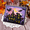 A square Halloween-themed card or art print featuring black haunted houses, bats, a witch, a ghost, and a crescent moon against a purple sky, displayed on a white lace doily over a rustic wooden surface with autumn leaves.