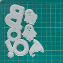 Five silver metal die-cut shapes for crafting, including a ghost, a pumpkin, a boot, and a letter 'o', arranged on a green cutting mat with grid lines.
