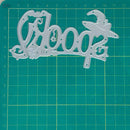 A silver metal die-cut craft stencil with the word 'hoop' in decorative script and a witch hat, placed on a green cutting mat with a yellow grid.