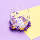 A clear glass candle holder with purple butterfly decals, holding a white tea light candle, set on a purple and yellow paper background with a purple flower base.