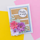 A handmade greeting card with a white border, light blue scalloped trim, a wood-grain panel, a circular white label reading 'Make a wish,' and layered paper flowers in pink, purple, and white with pale green leaves. The card is photographed on a colorful flat-lay background of yellow, pink, and white paper.