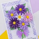 A handmade greeting card featuring layered paper flowers in shades of purple, pink, and white with green leaves and yellow button centers, accented by a purple paper bow at the bottom.