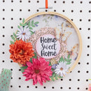 A decorative hanging hoop wall art piece with a wooden embroidery hoop, colorful paper flowers, butterflies, green leaves, and a central pink circle that reads “Home Sweet Home,” hung on a white pegboard.