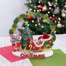 A festive Christmas tabletop decoration featuring a red sleigh with gold trim, a small green tree with a gold star, wrapped gift boxes, a green garland arch, and a white base that reads “Merry Christmas.” The scene is styled on a table with a blurred Christmas tree in the background.