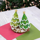 A handmade pop-up greeting card featuring two decorated Christmas tree pop-ups on a wood slice base, displayed on pink and green paper against a blurred festive background.