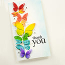 A handmade greeting card featuring a vertical arrangement of colorful paper butterflies in red, orange, yellow, green, blue, and purple on a light blue watercolor background, with the black script text 'thank you' on the lower right.