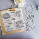 A clear stamp sheet with autumn-themed embossed designs and phrases, clipped to a kraft paper envelope, alongside matching metal stamp dies on a gray surface with a white flower prop.