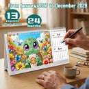 A spiral-bound desk calendar displayed on a wooden table, featuring a colorful turtle illustration on the front cover and monthly calendar pages on the right. A person's hand holds a black pen near the calendar, with a coffee cup, books, and glasses on the table.