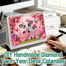 A DIY handmade diamond painting two-year desk calendar displayed on a desk. The calendar features a pink and white cow design with a bow, surrounded by pink roses and butterflies, with the word 'LOVE' spelled out in pink rhinestones.