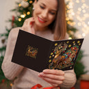 A woman holds an open black Christmas card featuring a detailed gold and multicolor reindeer illustration with stars and holiday decorations, with a blurred Christmas tree and warm lights in the background.