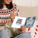 A person holding a white greeting card with a winter illustration, seated on a couch. The person wears a multicolor knit sweater and blue jeans, with a red-and-white striped candy cane visible nearby.