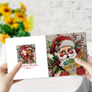 Two hands holding an open diamond painting kit showing Christmas-themed designs: a deer with red berries on the left page and Santa Claus with gifts on the right page, against a blurred background of yellow flowers and a white brick wall.