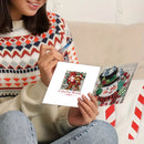 A person sitting on a couch holds a diamond painting canvas featuring a colorful Christmas ornament, with another completed snowman canvas beside it. The person wears a patterned knit sweater and blue jeans in a cozy indoor setting.