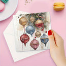 A hand with red nail polish holds a square greeting card featuring a watercolor design of hanging Christmas ornaments, with a white envelope behind it on a pink background.
