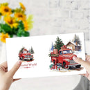 A person holds a white card or print featuring two watercolor-style red vintage trucks in winter scenes, with one small version and one larger decorated truck. The background is a softly blurred indoor setting with yellow flowers on the left and a light wall behind.
