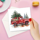 A hand with red nail polish holds a square greeting card featuring a watercolor illustration of a red pickup truck carrying a Christmas tree and wrapped gifts, set against a pink background with a white envelope and macarons.