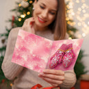 A woman in a cozy knit sweater holds an open pink holiday card featuring a pair of pink knit mittens with snowflake and rhinestone details, with a blurred Christmas tree and warm string lights in the background.