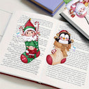 A close-up of an open book with printed text, decorated with colorful illustrated stickers or patches featuring a green-and-white elf, a penguin in a basket, and red strawberry motifs. A hand is visible at the top right, placing or adjusting one of the decorative pieces.