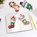 A flat-lay view of a diamond painting or craft kit showing Christmas-themed stocking designs. An open book displays two completed stockings, one with an elf and one with Santa, while two additional stocking designs are placed above on a white desk next to a yellow pencil.