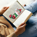 A person holding an open book with printed text, featuring a Christmas-themed pop-up page insert shaped like a green stocking with Santa and a child, styled on blue jeans and a beige sweater.
