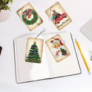 Four Christmas-themed diamond painting canvas tags are displayed on a white desk, including a wreath, a red truck with 'Merry Christmas', a Christmas tree, and Santa Claus. An open notebook and a yellow pencil are visible in the background as props.