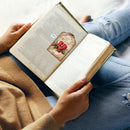A person holding an open book in their lap, wearing a tan sweater and blue ripped jeans, with a small Christmas-themed book feature visible on the left page.