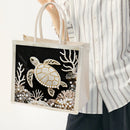 A person wearing a white and gray striped shirt holds a canvas tote bag decorated with a large sea turtle and coral designs made from white pearls and beads on a black background.