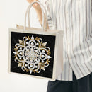A person wearing a white and gray striped shirt holds a beige canvas tote bag with a large black front panel featuring an intricate white and gold mandala-style floral pattern.