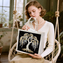 A woman seated in a hanging rope chair holds a white tote bag with a black front panel featuring a large rhinestone tree design. The setting is a softly lit indoor space with plants and window light in the background.