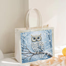 A white canvas tote bag with a large decorative winter owl print on the front, featuring a blue owl perched on a snowy branch with white snowflake details, photographed in a softly lit indoor setting.