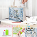 Pearl-decorated canvas diamond painting bag featuring an owl on a branch with snowflakes, shown with a hand placing a white sheet inside. The bag has white handles and is displayed on a textured blanket with a red pencil and paintbrushes nearby.