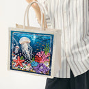A person wearing a white striped shirt holds a canvas tote bag with an underwater ocean scene design featuring a jellyfish, starfish, coral, and fish on a dark blue background.