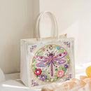 A clear plastic tote bag with white fabric handles, featuring a colorful beaded dragonfly and floral design on the front, set against a softly lit neutral background.