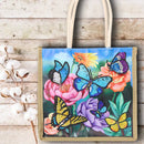 A canvas tote bag with a colorful printed design of blue, yellow, and purple butterflies on pink and purple flowers, set against a rustic light wood background with cotton bolls on the left.